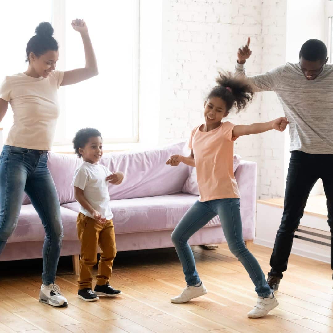 A happy Black family of four dancing together in their living room. The parents and two children smile and move with energy and joy, enjoying quality time during a lively family dance party.