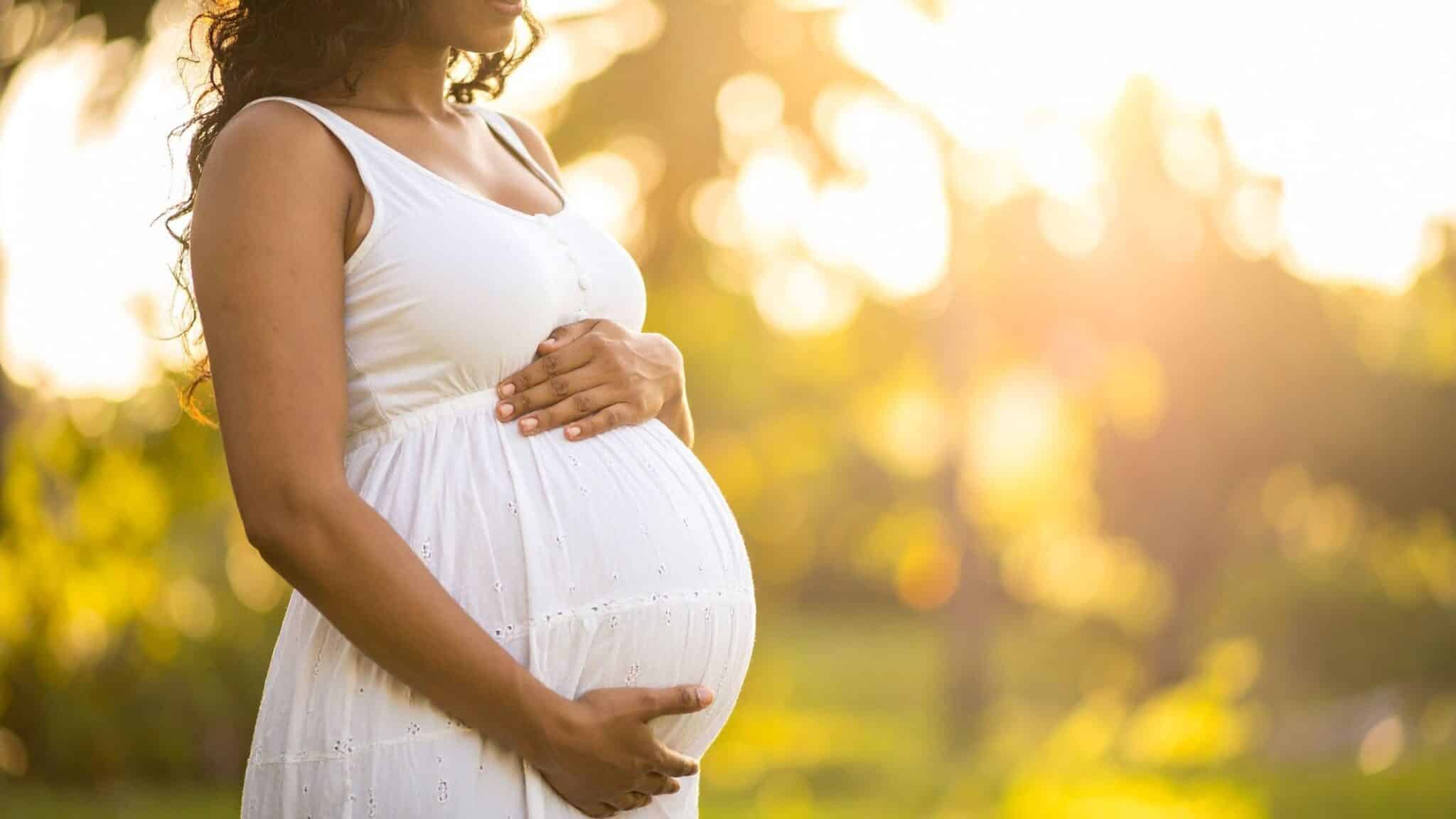 Pregnant woman in a white dress gently holding her baby bump outdoors at sunset, symbolizing healthy pregnancy and prenatal care in Madison WI.