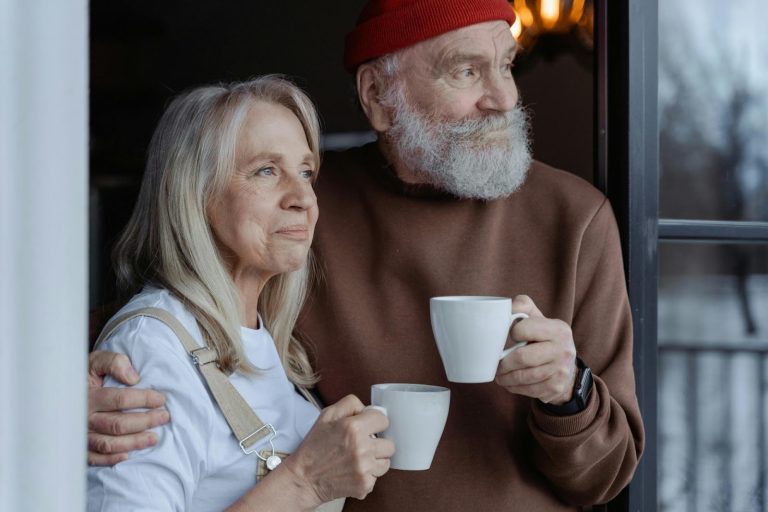 Older couple standing by window holding coffee mugs and looking ahead
