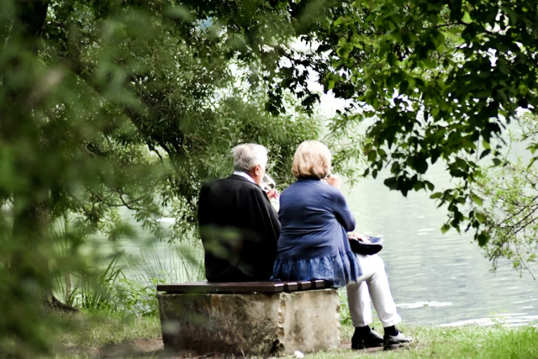 Elderly couple sitting together on a bench by a lake surrounded by trees