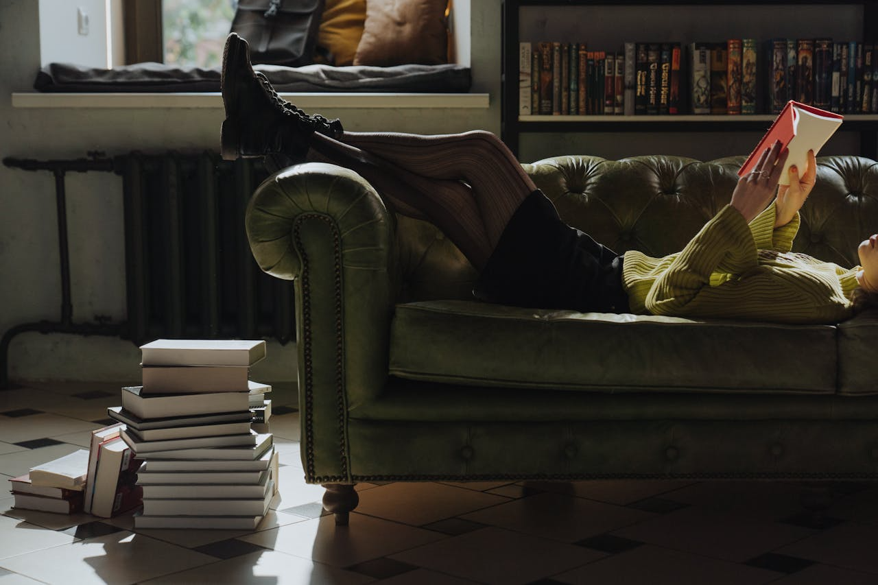 Woman relaxing on green sofa reading a book beside a stack of books