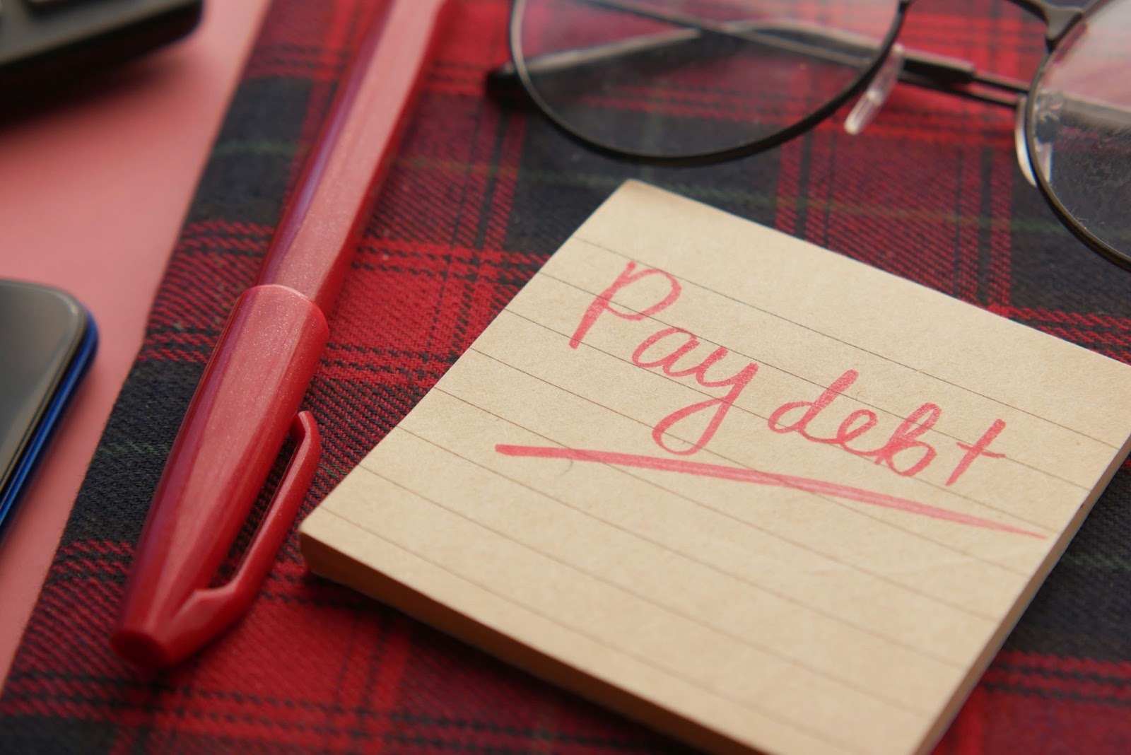 Sticky note that says pay debt with pen, glasses, and phone on a desk