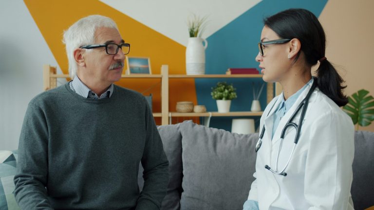 A female healthcare professional in a white coat sits on a couch, talking collaboratively with an older male patient about managing a chronic health condition.