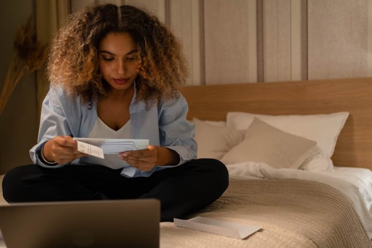 Woman sitting on bed reviewing utility bills and expenses at home