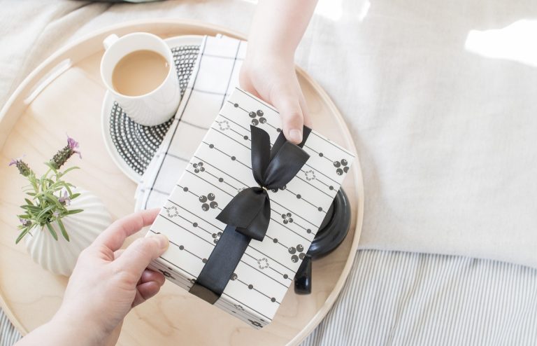 person handing a wrapped sweet treat gift on a tray with coffee and flowers