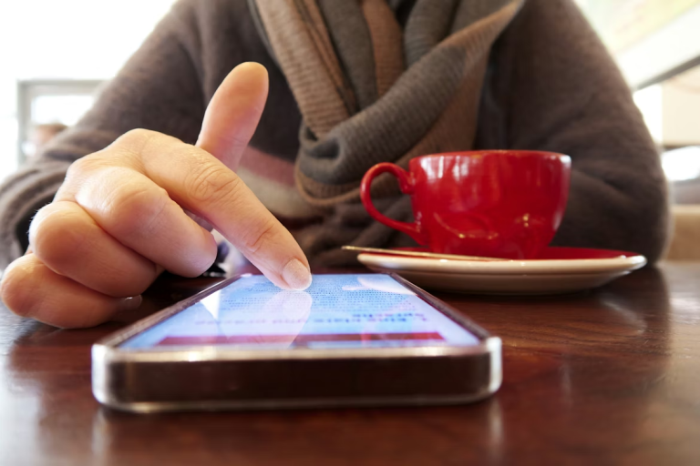 Person scrolling on a smartphone while sitting at a table with a cup of coffee.