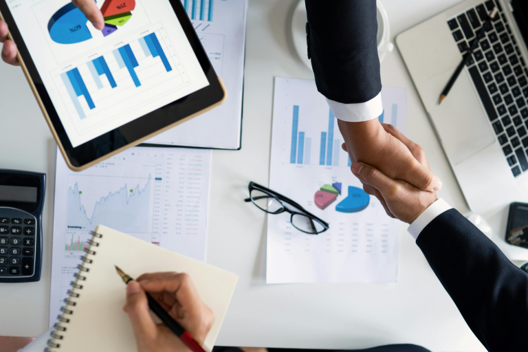 Two business professionals shaking hands over financial charts and business planning documents.
