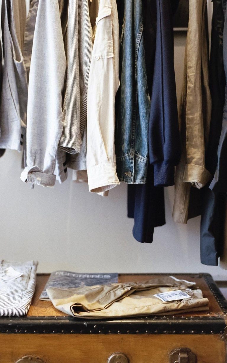Neutral-toned clothing hanging in a minimalist closet with folded clothes on a trunk below