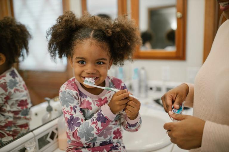 young girl brushing teeth with help from parent in bathroom
