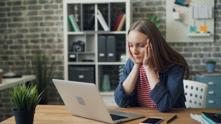 Woman sitting at a desk looking stressed while working on a laptop in a busy office