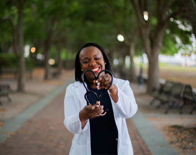 Smiling nurse holding a stethoscope in the shape of a heart while standing outdoors, symbolizing compassionate patient care and support.