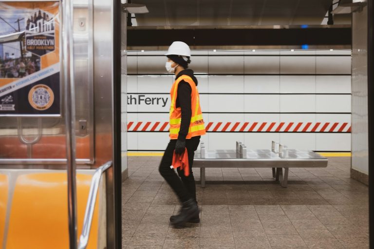 Employee wearing a safety vest, hard hat, and protective gear while working in a public transit station.