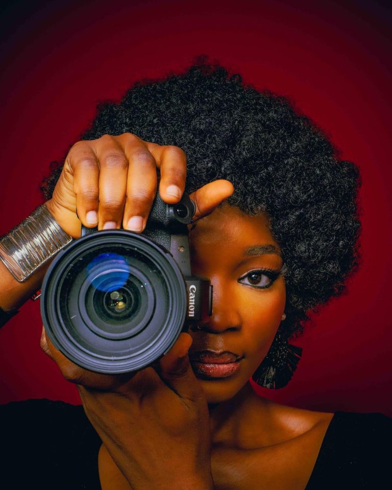 Female photographer holding a professional camera in front of her face against a bold red background