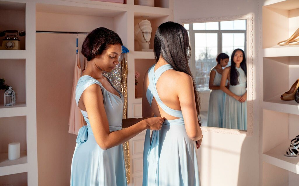 Bridesmaid fastening the back of another woman's dress while preparing for a wedding in a bridal suite.