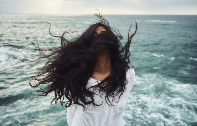 Woman with long dark hair standing by the ocean, symbolizing confidence, healing, and self-acceptance during a hair loss journey.
