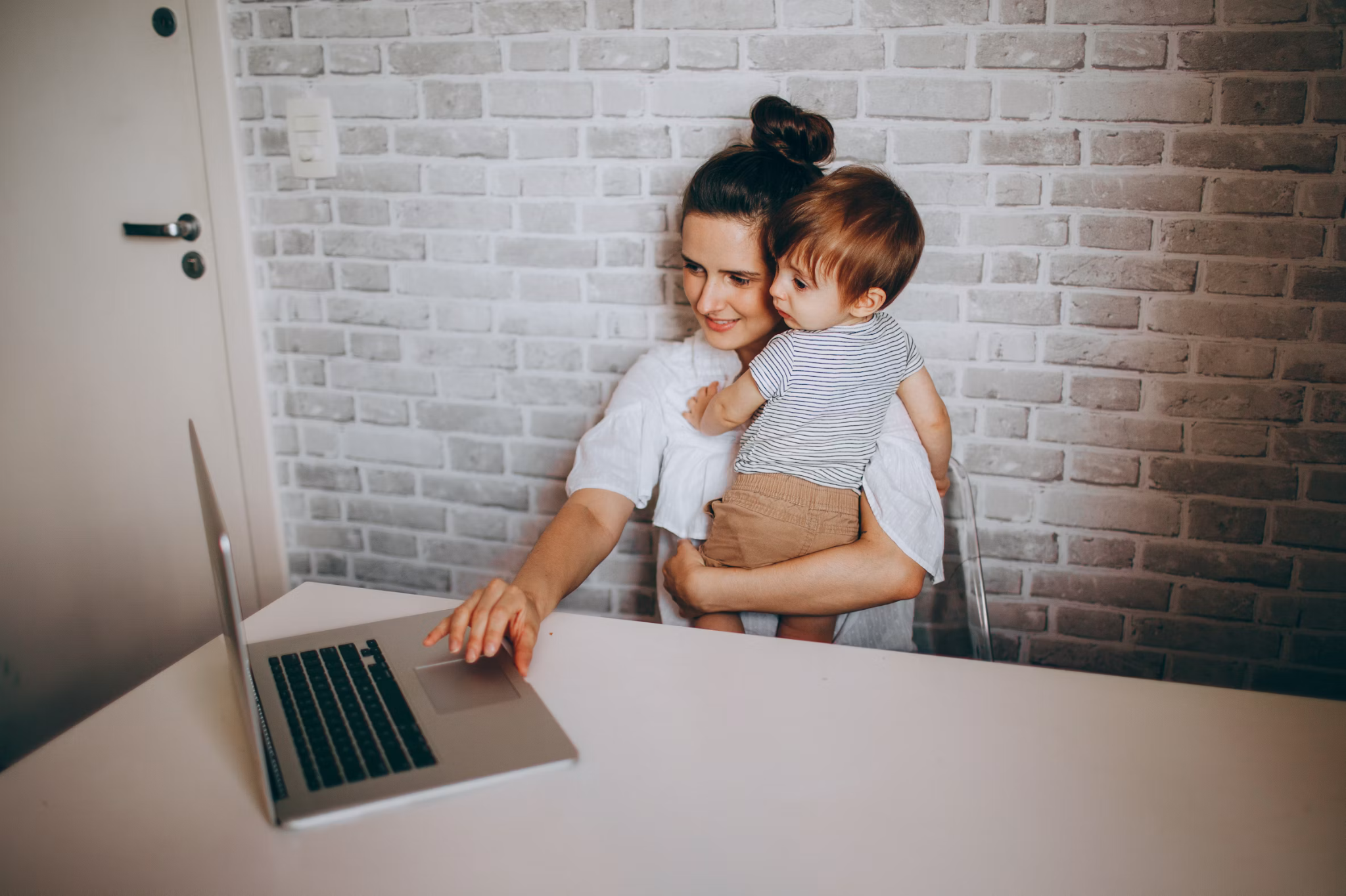 Mother holding her young child while working on a laptop as she prepares to return to work after maternity leave.
