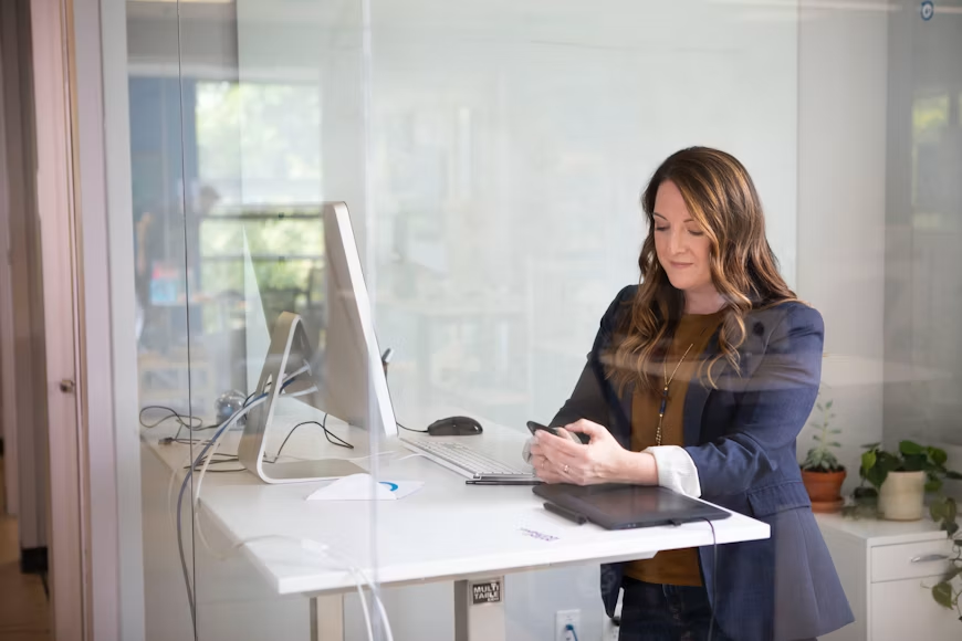 Business owner standing at a desk reviewing health insurance information on a smartphone in a modern office.