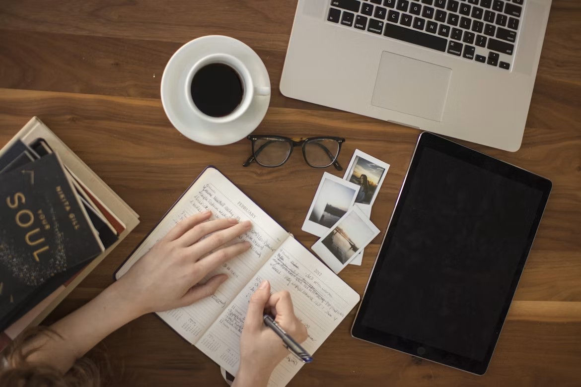 Person writing goals in a planner beside a laptop, tablet, books, and a cup of coffee.