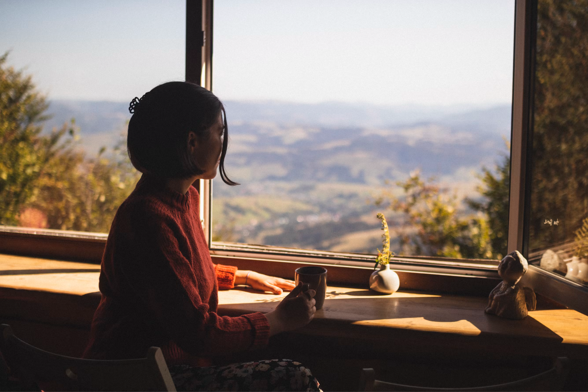 Woman sitting by a large window with a cup while reflecting on her life and priorities.