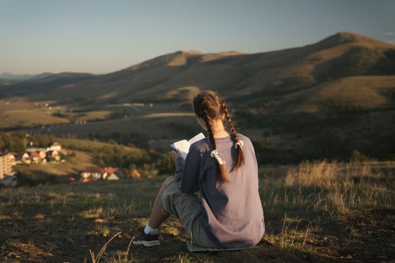 Woman reading a book outdoors while overlooking a peaceful mountain landscape