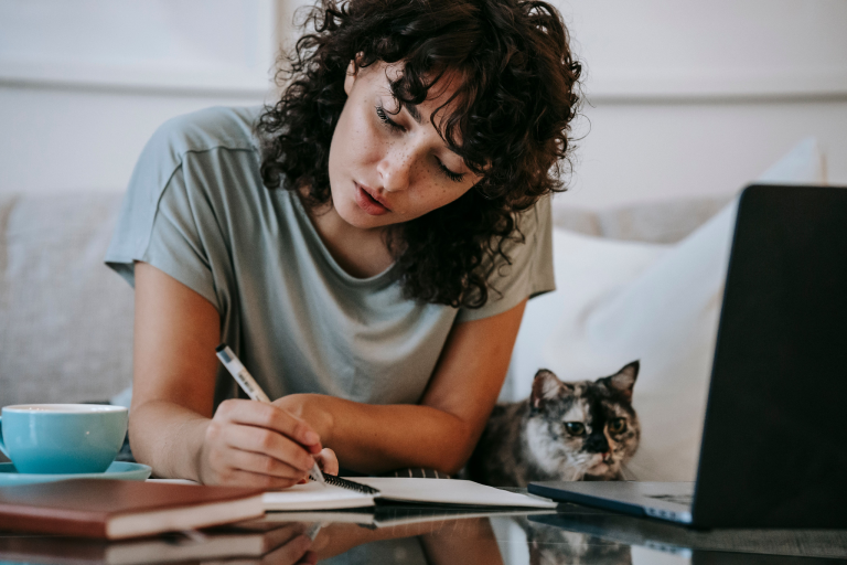 Woman writing wellness goals in a notebook beside a laptop at home