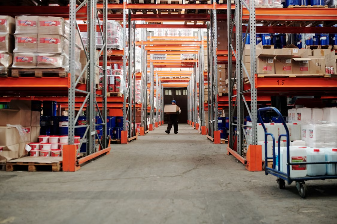 Warehouse worker carrying a box through an aisle between tall storage racks filled with inventory.
