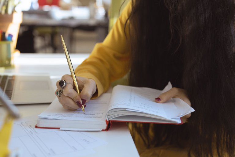 Woman writing in a planner beside a laptop and paperwork on a desk.