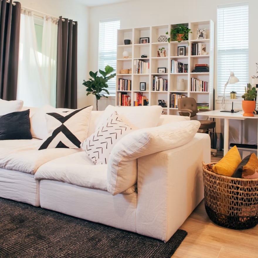 Bright, organized living room with a white sofa, bookshelf, desk, plants, and woven basket.