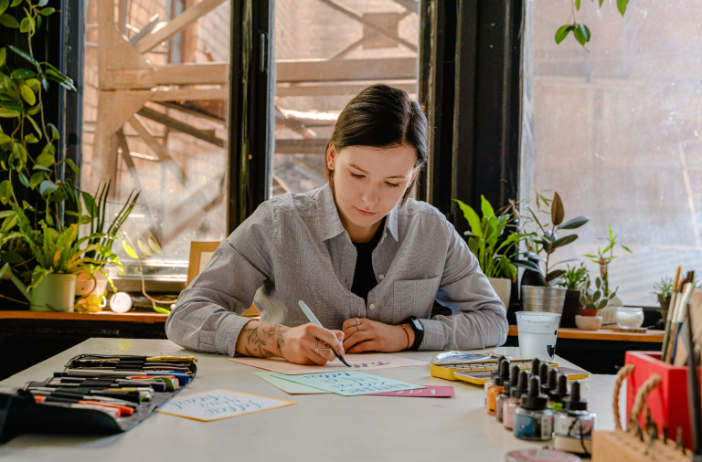 Woman creating colorful handwritten cards at a desk surrounded by art supplies and plants.