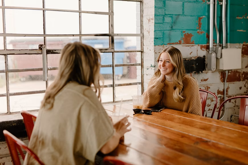 Two women having a relaxed conversation at a wooden table in a café.