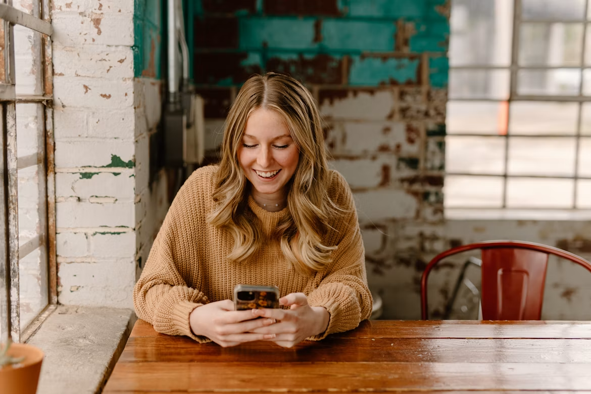 Woman smiling while using a smartphone at a wooden table in a café.