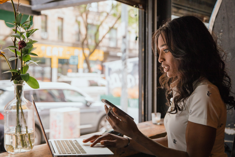 A woman using a laptop and smartphone at a table beside a window.