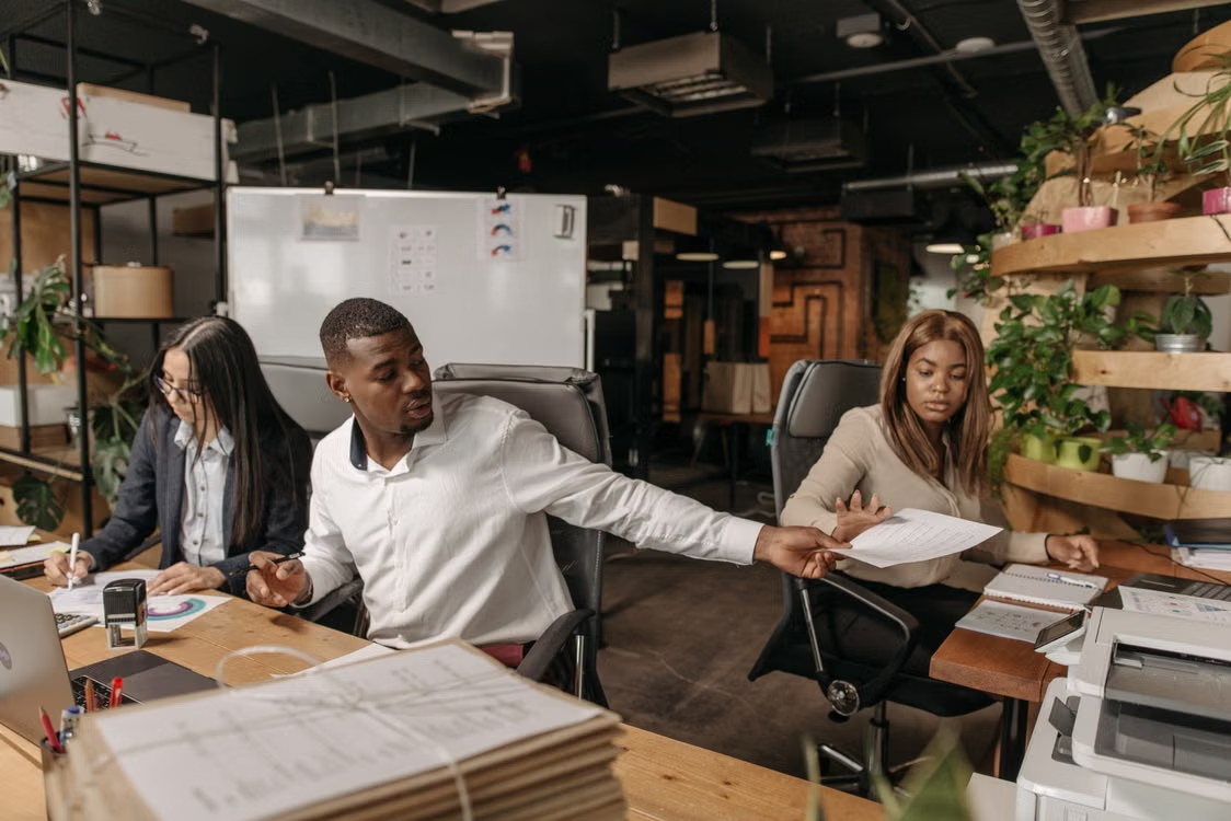 Three coworkers share documents and work at desks in a busy office.