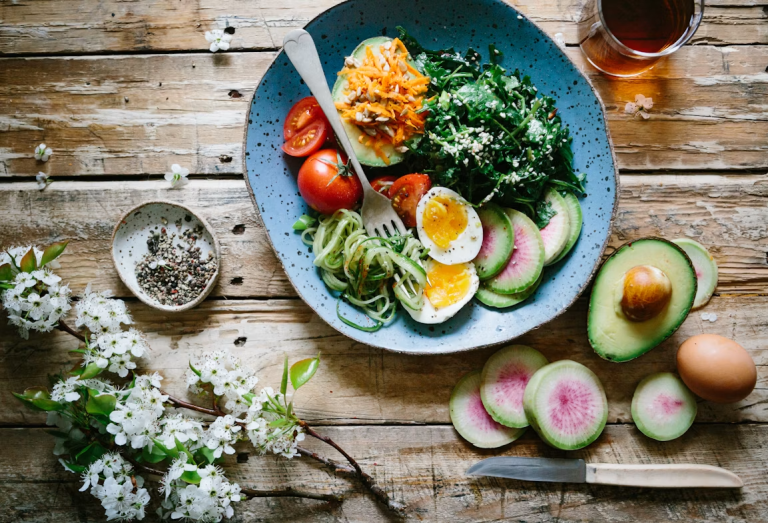 Colorful bowl with leafy greens, eggs, avocado, tomatoes, radishes, and shredded carrots on a wooden table.