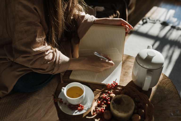 Person writing in a lined journal beside a cup of tea, teapot, and bowl on a wooden table.