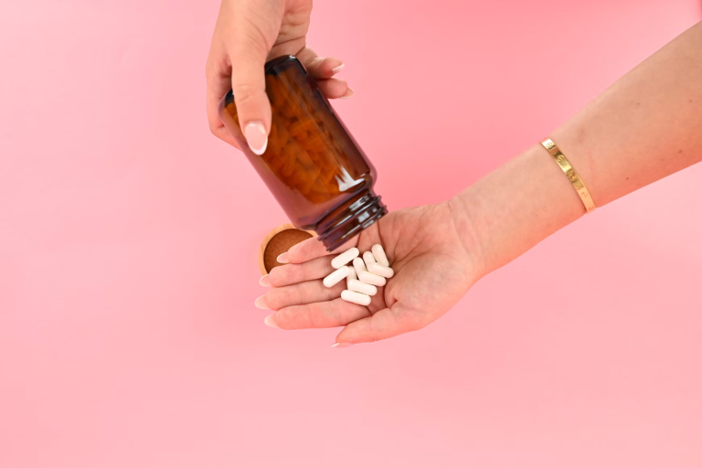 Hands pouring white capsules from an amber supplement bottle against a pink background.