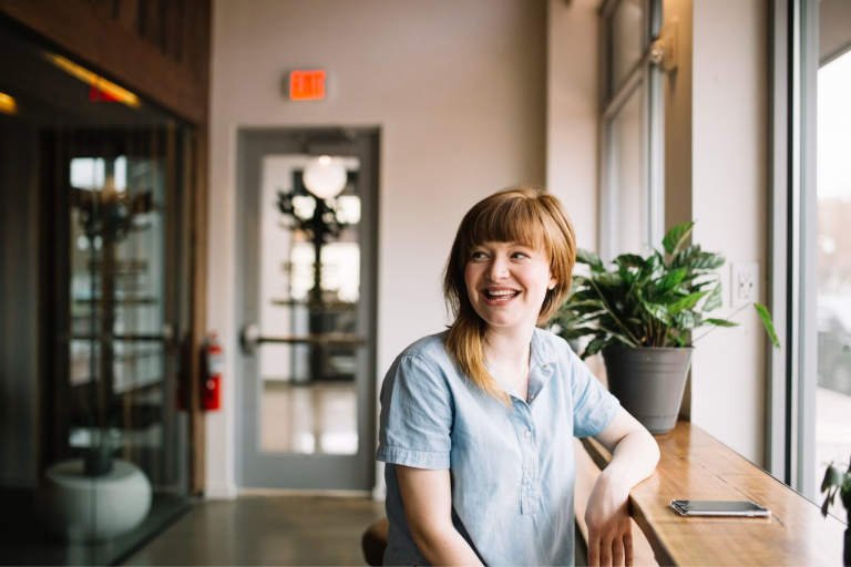 Smiling woman in a light blue top sitting by a window in a bright modern workspace.