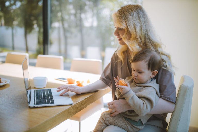 Woman working on a laptop while holding a toddler at a kitchen table.