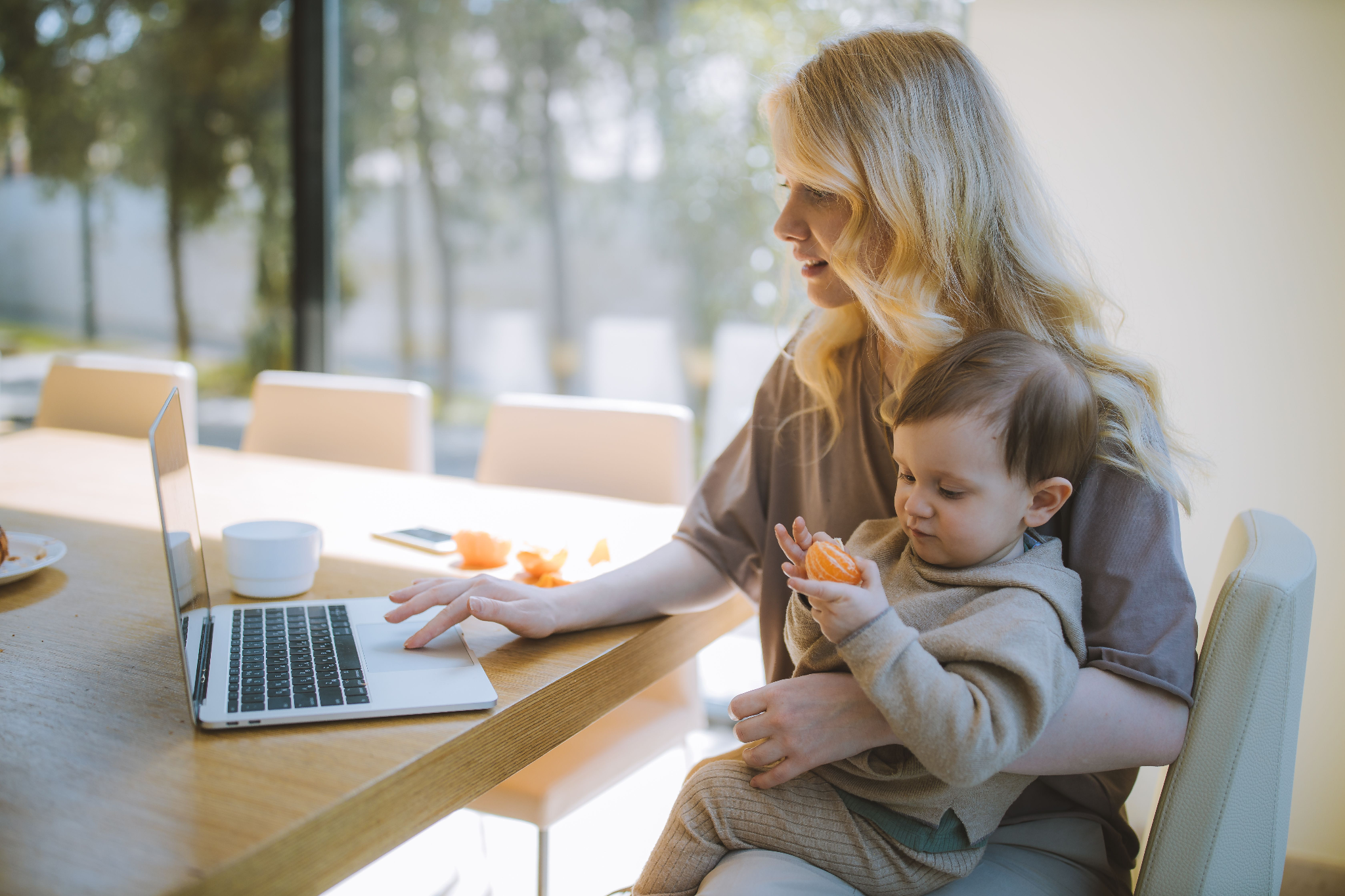 Woman working on a laptop while holding a toddler at a kitchen table.