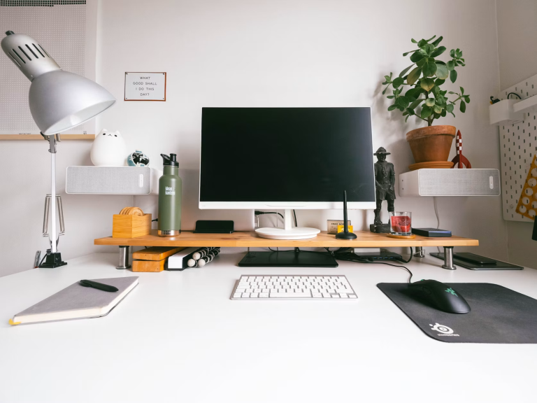 Organized home office desk with a computer monitor, keyboard, notebook, lamp, water bottle, and potted plant.