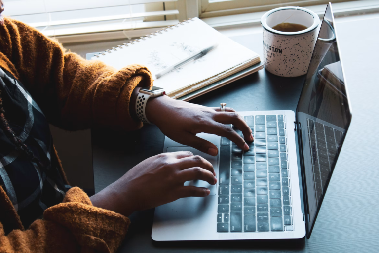 Person typing on a laptop beside an open notebook and a cup of tea at a desk.