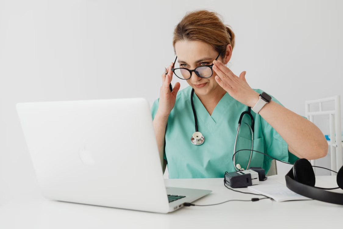 Healthcare worker wearing glasses adjusts them while looking at a laptop.