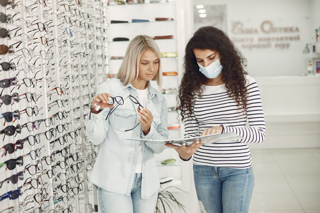 Two women review eyeglass frames at an optician’s shop beside a wall of glasses.