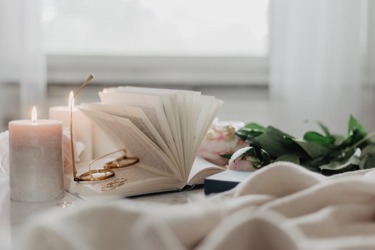 Open book, lit candle, eyeglasses, and green leaves arranged on a soft bed in a calm home setting.