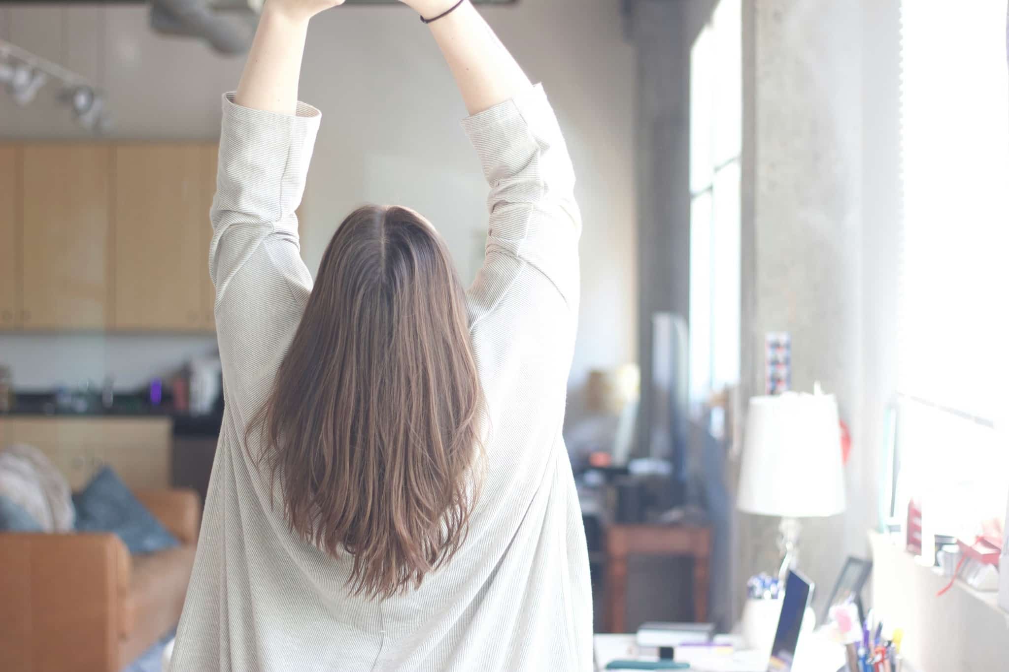Woman stretching with both arms raised near a sunny window in a home workspace.