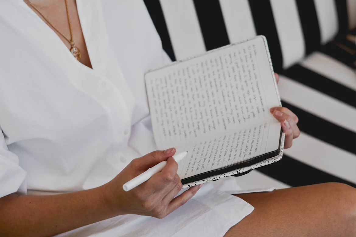 Woman writing in an open notebook with a white pen while seated on a black-and-white striped chair.