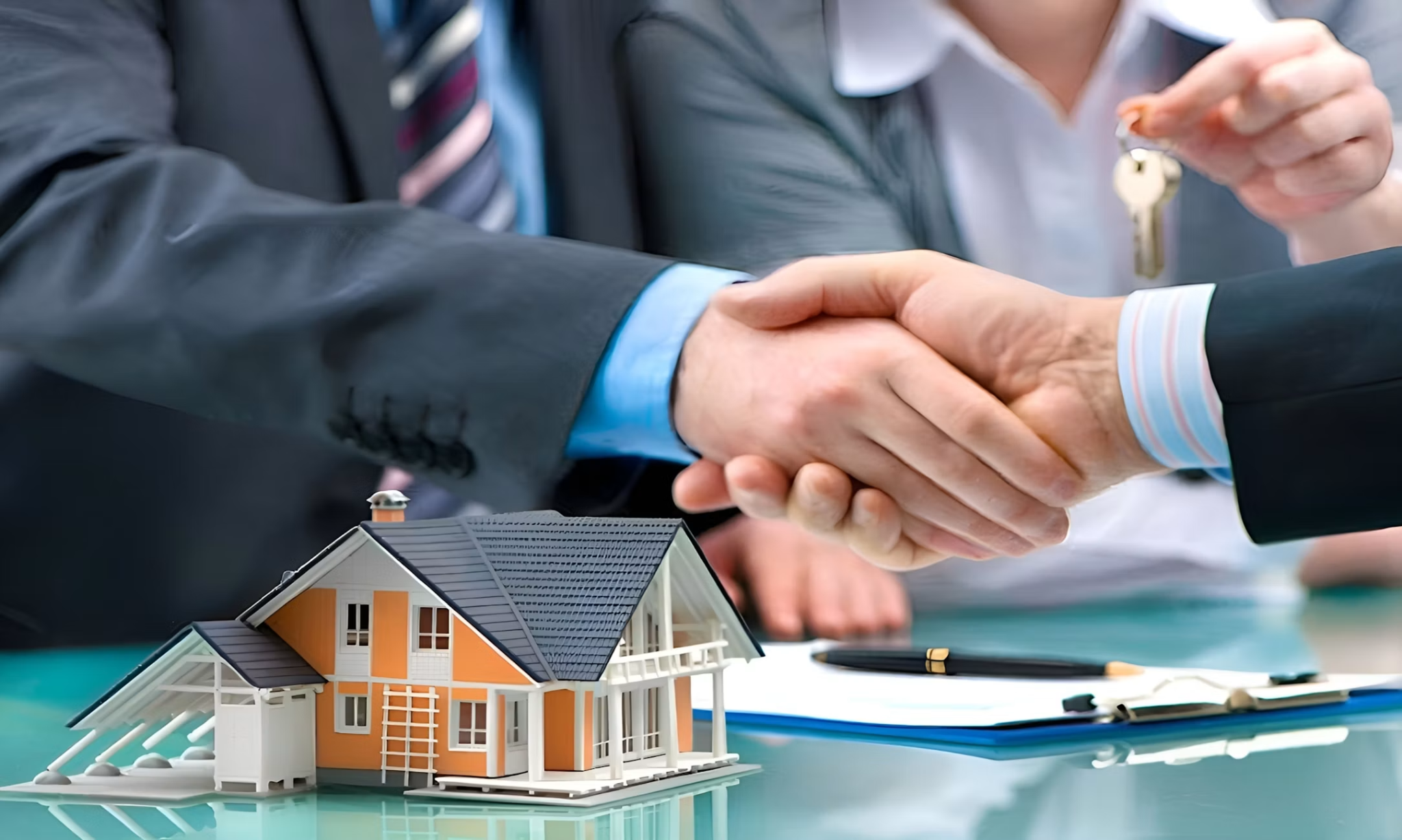 Two people shake hands over a model house while another person holds a set of keys.