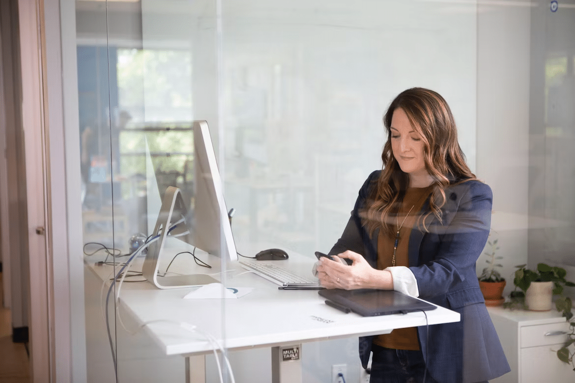 Woman entrepreneur working at a standing desk and reviewing her phone beside a computer and tablet.
