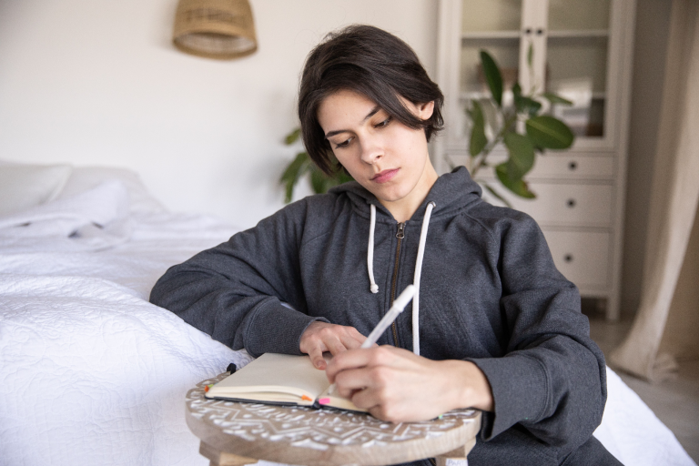 Woman sitting beside a bed and writing in a notebook to reflect on her goals and thoughts.