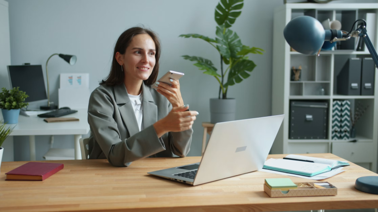 Woman at a home office desk using a smartphone beside an open laptop